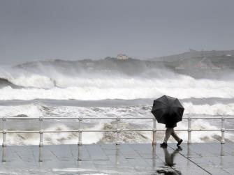 Person with umbrella walking on wet boardwalk in front of a raging ocean