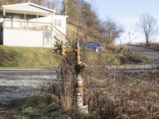 A rusty pipe sticking out of the ground with a house and car in the near background.